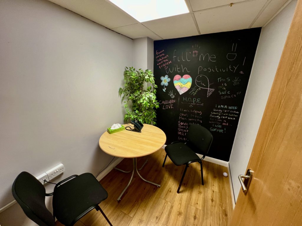 the photo shows an inside room from the Stevenage Crisis Cafe, including chairs and a table, a plant, plus a black wall with chalk artwork and writing