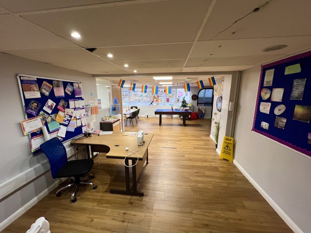 the photo shows the inside of our Stevenage Crisis cafe, which includes a desk and chair, rainbow flags hanging from the ceiling, two noticeboards with posters, a table and chairs, a water fountain, and a table football