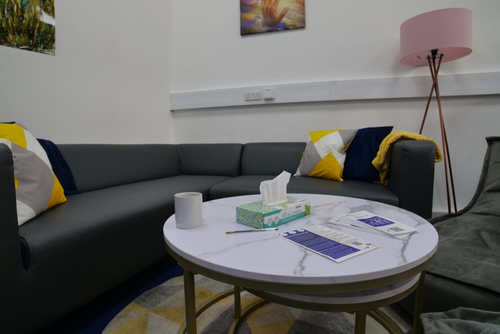 the photo shows an inside room from our Hatfield Crisis Cafe, which includes a long sofa and cushions, a table with leaflets and flyers, a lamp, art on the walls